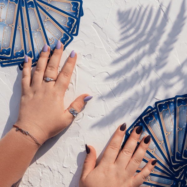 hands-with-purple-nails-rings-deck-tarot-cards-fanned-out-white-surface-with-fern-leaf-shadow-top-view